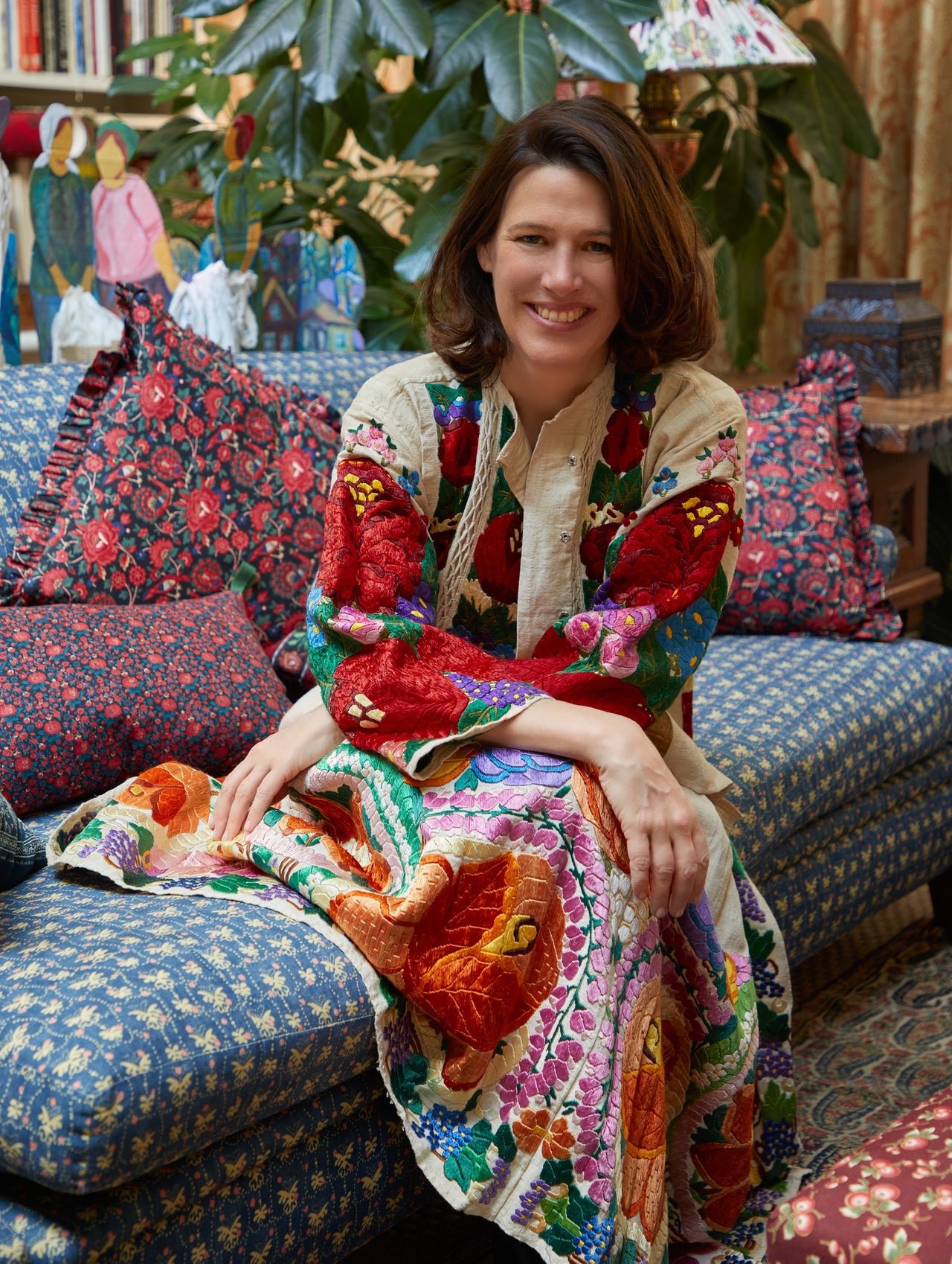 Nathalie Farman-Farma in a colorful embroidered dress sitting on a patterned couch with floral decorations in the background.