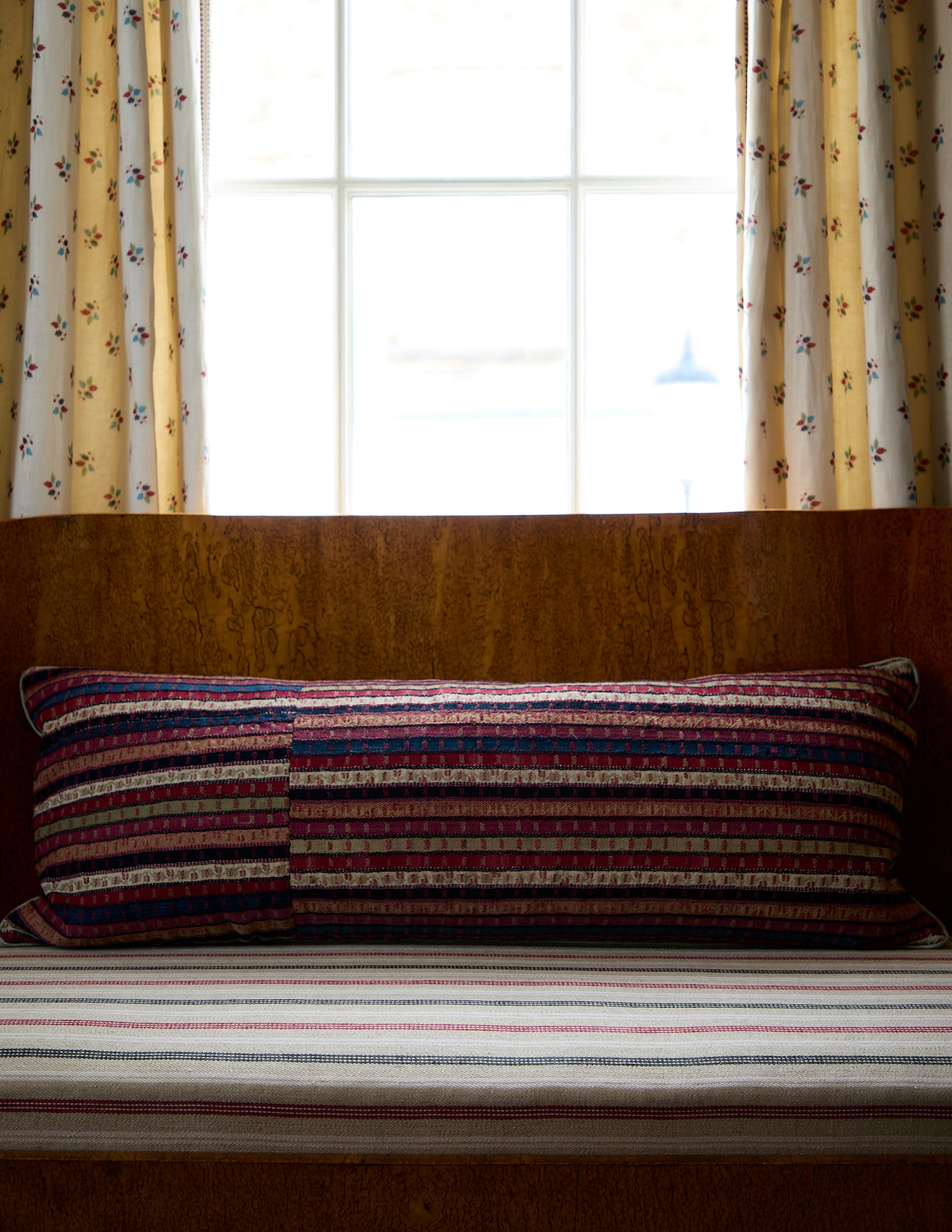 Striped pillow on a bench with Cara Delevingne fabric curtains and a window in the background