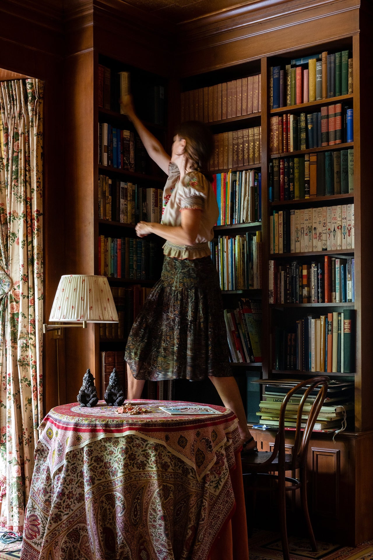 Décors Barbares founder, Nathalie-Farman-Farma standing on a chair in a room filled with bookshelves and Koubla fabric curtains.
